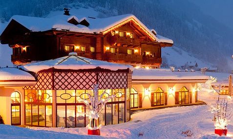 Ein großes Hotel im Chalet-Stil mit warmen Lichtern leuchtet in der Abenddämmerung in einer verschneiten Berglandschaft.