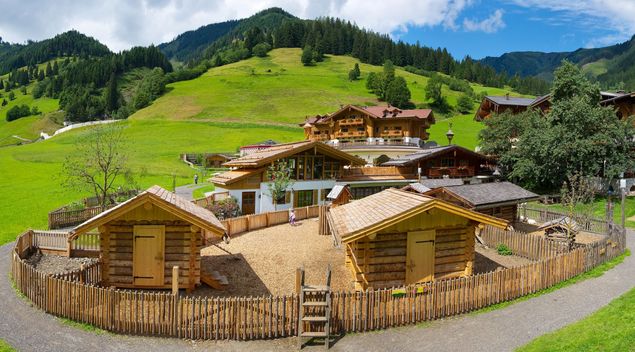 Ein eingezäuntes Gelände mit kleinen Holzhütten liegt zwischen größeren Gebäuden im alpinen Stil und grünen Hügeln unter einem teilweise bewölkten Himmel.