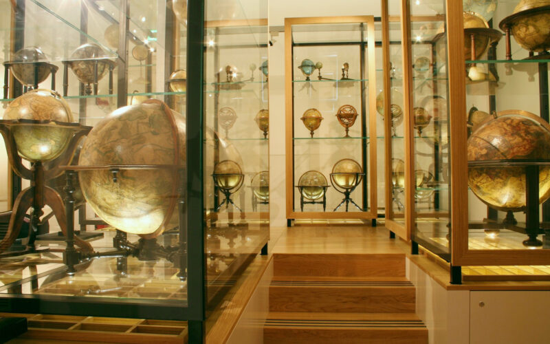 Eine museale Ausstellung antiker Globen in Glasvitrinen auf Holzpodesten, die verschiedene Größen und historische Kartendesigns zeigen.