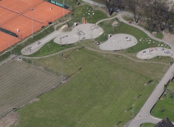 Luftaufnahme eines Parks mit einem Skatepark, Spielplatz, Bänken, Tennisplätzen und von Gras und Bäumen gesäumten Wegen.
