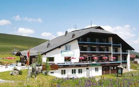 Ein Hotel im Chalet-Stil mit Balkonen und roten Sonnenschirmen auf einer Terrasse, eingebettet in eine grasbewachsene, hügelige Landschaft unter blauem Himmel.