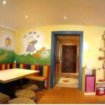 Buntes Kinderzimmer mit Wandmalereien, einer eingebauten Eckbank, kleinen runden Hockern, einem Bücherregal und leuchtend gelben Wänden.