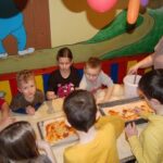 Eine Gruppe von Kindern sitzt um einen Tisch herum und bestreicht den Teig mit Soße, während ein Erwachsener in einem farbenfrohen Raum mit Wandmalereien und Luftballons Pizzen herstellt.
