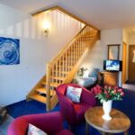 Ein gemütliches Hotelzimmer mit einem Kamin, roten Sesseln, einem Couchtisch mit Blumen, einem Fernseher, einem Schreibtisch, einer Treppe zu einem Loft und einem Bademantel, der am Eingang hängt.