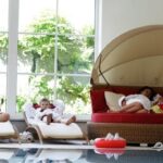 Eine Familie in weißen Bademänteln entspannt sich am Innenpool, mit Kindern auf Liegen und Erwachsenen, die den Kindern auf einer runden Liege in der Nähe eines großen Fensters vorlesen.