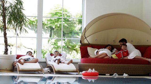 Eine Familie in weißen Bademänteln entspannt sich am Innenpool, mit Kindern auf Liegen und Erwachsenen, die den Kindern auf einer runden Liege in der Nähe eines großen Fensters vorlesen.