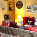 Gemütliches Wohnzimmer mit Korbmöbeln, roten und gemusterten Kissen, einer beleuchteten Lampe, Wandkunst und gelb gestrichenen Wänden.