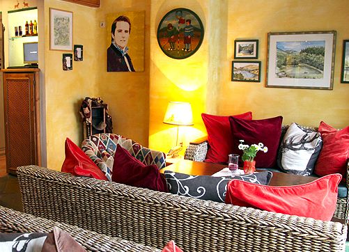 Gemütliches Wohnzimmer mit Korbmöbeln, roten und gemusterten Kissen, einer beleuchteten Lampe, Wandkunst und gelb gestrichenen Wänden.