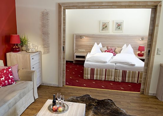 Moderne Hotelsuite mit einem Wohnbereich mit Sofa und Couchtisch und einem Schlafzimmer mit zwei Einzelbetten, roten Akzenten und dekorativen Kissen.