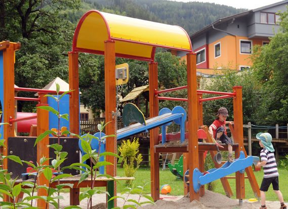 Zwei Kinder spielen auf einem farbenfrohen Spielplatz im Freien mit Rutschen und Klettergerüsten in einer Grünanlage mit Bäumen und nahe gelegenen Gebäuden.