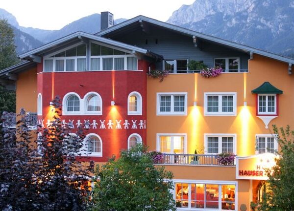 Ein farbenfrohes Hotel mit orangefarbenen und roten Wänden, vielen Fenstern, Balkonen mit Blumen und Außenbeleuchtung, eingebettet in eine bergige Landschaft.