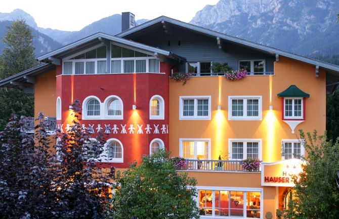 Ein farbenfrohes Hotel mit orangefarbenen und roten Wänden, vielen Fenstern, Balkonen mit Blumen und Außenbeleuchtung, eingebettet in eine bergige Landschaft.