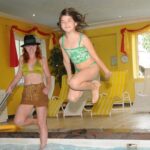 Ein Mädchen in einem grünen Badeanzug springt in ein Hallenbad, während eine Frau in der Nähe steht, mit gelben Liegestühlen und hellgelben Wänden im Hintergrund.