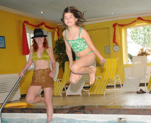Ein Mädchen in einem grünen Badeanzug springt in ein Hallenbad, während eine Frau in der Nähe steht, mit gelben Liegestühlen und hellgelben Wänden im Hintergrund.