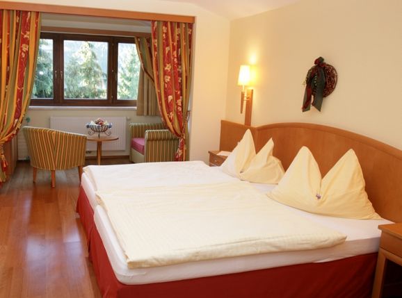 Ein Hotelzimmer mit einem Doppelbett, zwei Stühlen, einem kleinen runden Tisch am Fenster, Holzfußboden und roten und gelben Dekorationsakzenten.