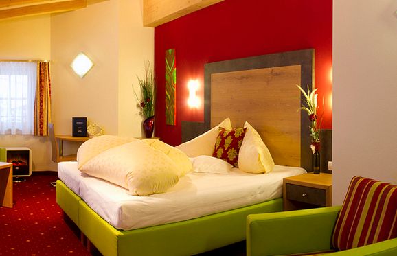 Hotelzimmer mit grünem Bett, weißer Bettwäsche, roter Akzentwand, moderner Einrichtung, zwei Nachttischen, einem Schreibtisch und einem Fenster mit durchsichtigen Vorhängen, die natürliches Licht hereinlassen.