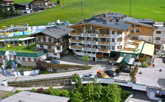 Ein mehrstöckiger Hotelkomplex mit Balkonen, umgeben von grünen Feldern und anderen Gebäuden, mit vor der Tür geparkten Autos und einem Poolbereich auf der linken Seite.