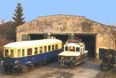 Zwei alte Triebwagen, einer blau und cremefarben, der andere braun und weiß, stehen vor einem alten Wartungsgebäude mit offenen Türen unter freiem Himmel.