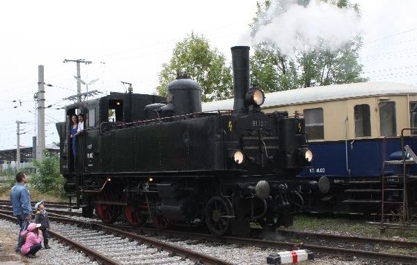 Eine schwarze Dampflokomotive mit beleuchteten Scheinwerfern stößt Dampf aus, während sie auf den Gleisen neben einem blau-beigen Personenwagen steht. Mehrere Personen sind in der Nähe und beobachten den Zug.
