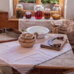 Eine rustikale Küche mit einem Holztisch, der mit Schüsseln, einem Schneidewerkzeug und einer Schüssel gedeckt ist; Gläser mit Eingemachtem und Getreidesäcke stehen in der Nähe eines sonnendurchfluteten Fensters.