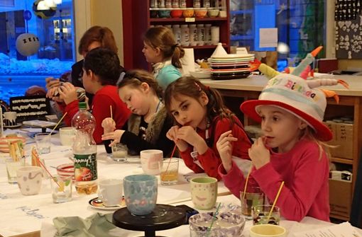Kinder sitzen an einem Tisch und bemalen Keramikbecher und -schalen; ein Mädchen trägt einen bunten Geburtstagshut. Auf dem Tisch stehen Malutensilien und Getränke. Erwachsene beaufsichtigen im Hintergrund.