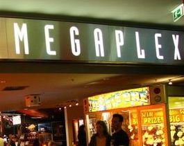 Ein beleuchtetes „MEGAPLEX“-Schild über dem Eingang zu einem Unterhaltungs- oder Kinolokal, darunter stehen Menschen und im Hintergrund sind Spielautomaten zu sehen.