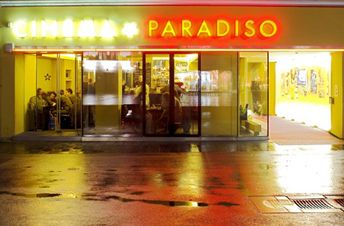 Der neonbeleuchtete Eingang des Cinema Paradiso bei Nacht, mit Menschen, die durch die Glastüren ins Innere schauen, und Spiegelungen auf der nassen Straße draußen.