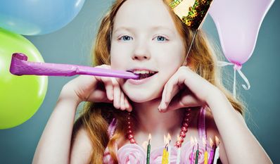 Junges Mädchen mit roten Haaren, das einen Partyhut trägt und in ein Partyhorn bläst, sitzt vor einer Geburtstagstorte mit brennenden Kerzen und ist von Luftballons umgeben.