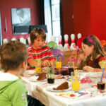Kinder sitzen um einen geschmückten Tisch herum, essen Kuchen und trinken Saft auf einer Geburtstagsfeier, mit Luftballons und Bowlingkegeln im Hintergrund.