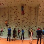 Eine Gruppe von Personen in einer Sporthalle, in der mehrere Personen eine große Kletterwand erklimmen, während andere zusehen oder warten, bis sie an der Reihe sind.