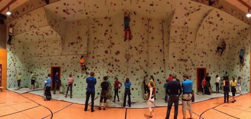 Eine Gruppe von Personen in einer Sporthalle, in der mehrere Personen eine große Kletterwand erklimmen, während andere zusehen oder warten, bis sie an der Reihe sind.