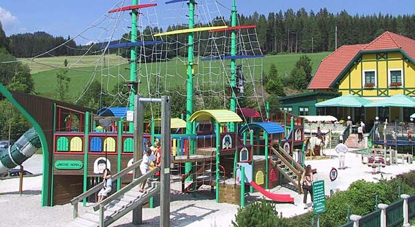Spielplatz im Freien mit einem großen Piratenschiff mit Rutschen, Kletternetzen und spielenden Kindern vor einer Kulisse aus Bäumen und einem Gebäude mit rotem Dach.