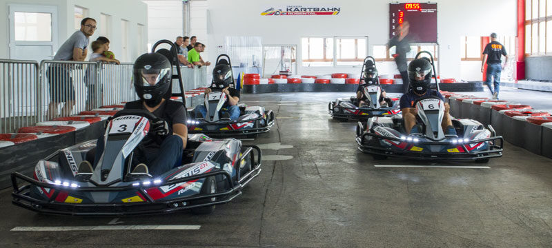 Drei Personen mit Helmen fahren Go-Karts auf einer Indoor-Rennstrecke, während Zuschauer hinter einer Absperrung zuschauen. Im Hintergrund ist eine digitale Zeitmessanlage zu sehen.