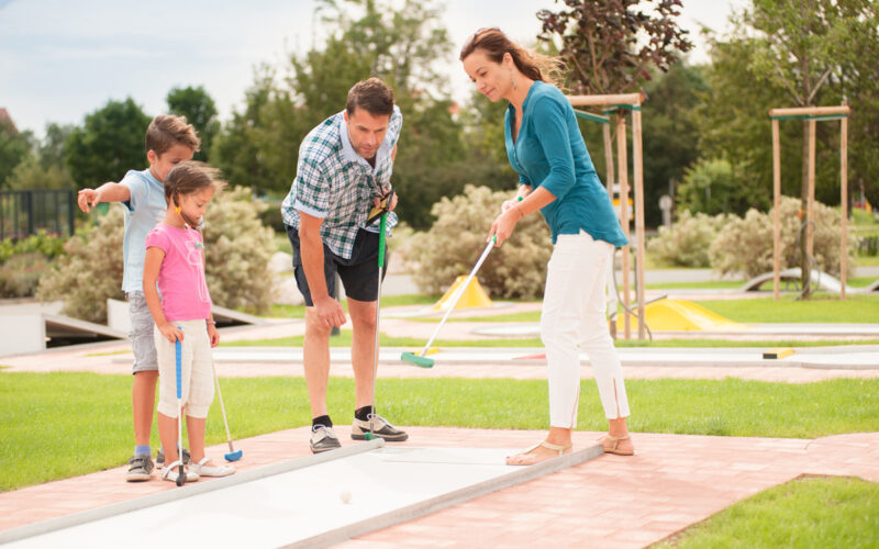 Eine vierköpfige Familie spielt im Freien Minigolf; die Eltern und zwei Kinder stehen mit Golfschlägern in der Hand auf dem Platz und konzentrieren sich auf das Spiel.