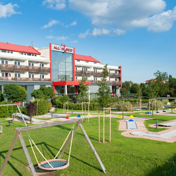 Ein Hotel mit rotem Dach und Balkonen überblickt an einem sonnigen Tag einen angelegten Spielplatz und eine Minigolfanlage.