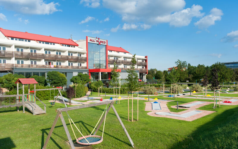 Ein Hotel mit rotem Dach und Balkonen überblickt an einem sonnigen Tag einen angelegten Spielplatz und eine Minigolfanlage.