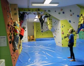 Mehrere Kinder klettern an einer Indoor-Boulderwand, während eine Person auf dem gepolsterten Boden steht und sie beobachtet.