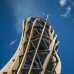 Ein hoher, spiralförmiger hölzerner Aussichtsturm mit Metallstützen steht vor einem klaren blauen Himmel mit ein paar Wolken.