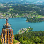 Von einem hohen hölzernen Aussichtsturm aus überblickt man einen See, der von bewaldeten Hügeln, kleinen Städten und grünen Feldern umgeben ist.