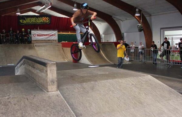 Ein BMX-Fahrer führt in einem Indoor-Skatepark einen Trick in der Luft aus, während die Zuschauer hinter einem Geländer zuschauen.