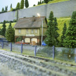 Eine detaillierte Modelleisenbahnszene mit einem kleinen Gebäude, Bäumen, Fahrzeugen, Eisenbahnschienen und einem Zaun im Vordergrund.