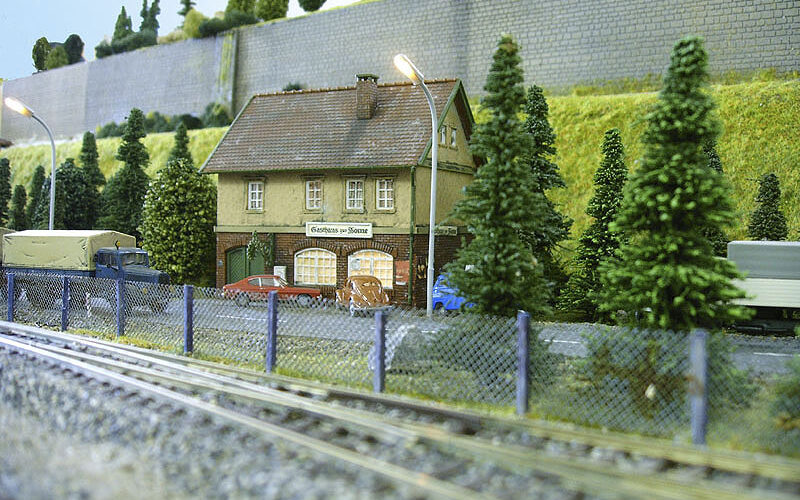 Eine detaillierte Modelleisenbahnszene mit einem kleinen Gebäude, Bäumen, Fahrzeugen, Eisenbahnschienen und einem Zaun im Vordergrund.