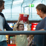 Ein Kind in einem gestreiften Hemd hält einen großen Popcorn-Eimer auf dem Kopf, während zwei Erwachsene das Kind in einem modernen Gebäude anlächeln.