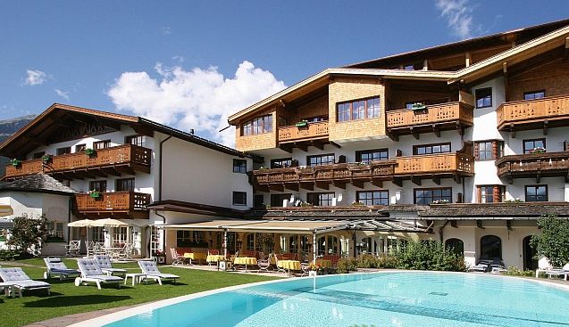 Ein Hotel mit Holzbalkonen und einem schrägen Dach steht hinter einem klaren Außenpool und Liegestühlen an einem sonnigen Tag.