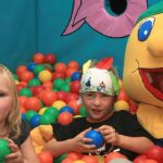 Drei Kinder sitzen in einem bunten Bällebad, eines trägt ein Stirnband mit Zeichnungen, ein anderes ein Figurenkostüm mit einem großen lächelnden Gesicht.