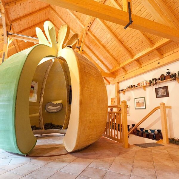 Eine große, hohle Skulptur in Form eines Apfels steht in einem Raum mit Holzbalken, gefliestem Boden und Regalen mit verschiedenen Gegenständen.