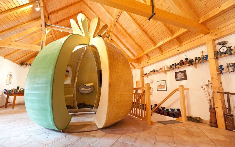 Eine große, hohle Skulptur in Form eines Apfels steht in einem Raum mit Holzbalken, gefliestem Boden und Regalen mit verschiedenen Gegenständen.