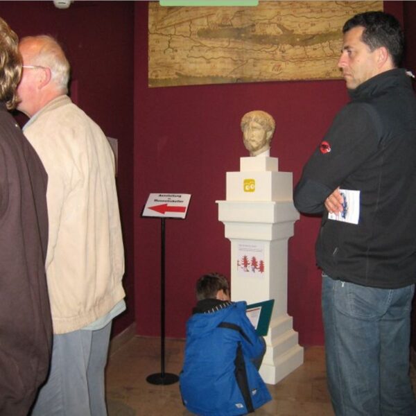 Vier Personen in einem Museumsraum mit roten Wänden; drei Erwachsene stehen neben einer Statuenbüste, während ein Kind in einer blauen Jacke sitzt und auf einem Klemmbrett schreibt oder zeichnet.