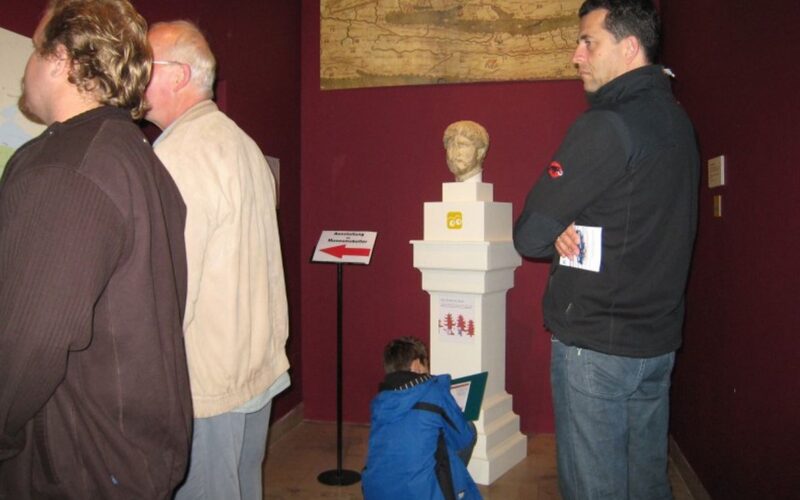 Vier Personen in einem Museumsraum mit roten Wänden; drei Erwachsene stehen neben einer Statuenbüste, während ein Kind in einer blauen Jacke sitzt und auf einem Klemmbrett schreibt oder zeichnet.