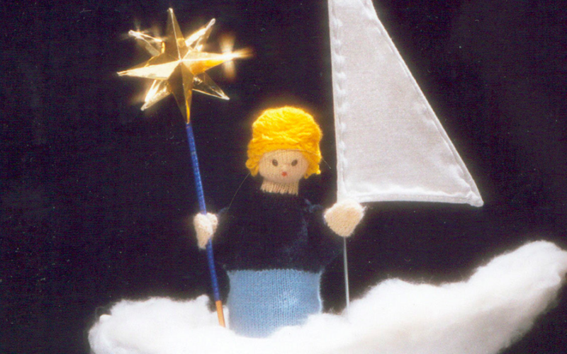 Eine handgefertigte Puppe mit gelbem Haar steht auf einer weißen, flauschigen Oberfläche und hält einen Sternenstab und eine weiße Fahne vor einem dunklen Hintergrund.